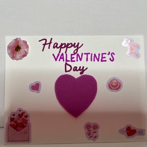 May include: A white card with the text "Happy Valentine's Day" written in purple. The card is decorated with pink hearts, flowers, and a large purple heart.