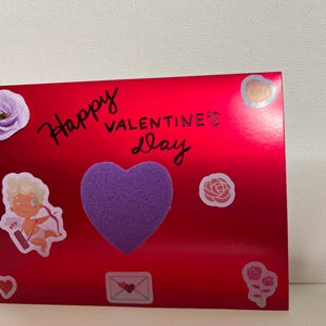May include: Red card with the text "Happy Valentine's Day" written in black marker. The card has a purple heart, a pink rose, a pink envelope, a pink flower, and a cartoon cupid with an arrow.