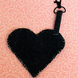 May include: A black heart-shaped keychain charm with a black metal clasp and strap. The charm has a textured surface and is set against a pink background with a white floral pattern.