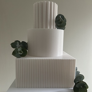 Puede incluir: Un pastel de boda blanco de tres pisos con un piso superior texturizado y acanalado y un piso inferior liso. El pastel está decorado con ramitas de eucalipto verde.