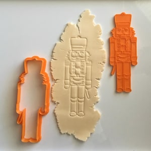 Può includere: Due stampini per biscotti in plastica arancione a forma di schiaccianoci. Uno stampino è pieno e l'altro è un contorno. Lo stampino pieno è su un pezzo di pasta per biscotti bianca.