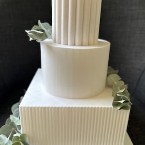 Może przedstawiać: Trzypiętrowy biały tort o teksturowanym wzorze. Dolne piętro jest kwadratowe, środkowe okrągłe, a górne cylindryczne. Zieleń akcentuje ciasto. Nowoczesny i elegancki design na wesele lub specjalną okazję.