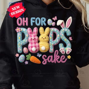 以下が含まれることがあります： ピンク、青、白で「OH FOR PEEPS SAKE」と書かれた黒いパーカー。イースターバニー、卵、ニンジン、蝶のデザイン。左上に「NEW DESIGN」のラベルがあります。