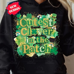 May include: Black hoodie with a St. Patrick's Day design. The design features the text "Cutest Clover in the Patch" in green and gold, along with shamrocks, rainbows, and a pot of gold. A "New Design" label is in the upper left.