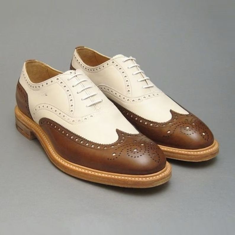 May include: A pair of brown and white leather brogue shoes with a wingtip design. The shoes have a lace-up closure and a brown leather sole.