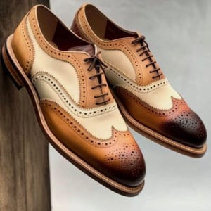 May include: A pair of brown, tan, and white leather oxford shoes with wingtip detailing and lace-up closures.