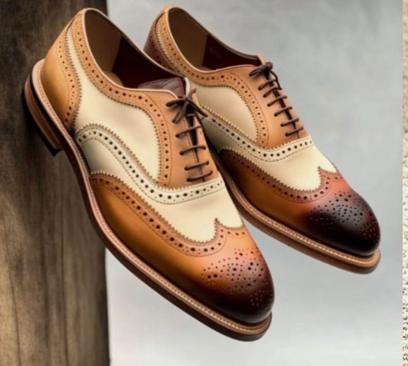 May include: A pair of brown, tan, and white leather oxford shoes with wingtip detailing and lace-up closures.