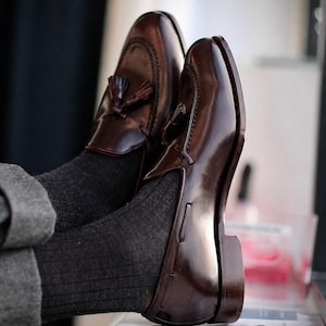 Puede incluir: Un par de mocasines de cuero marr&oacute;n oscuro con borlas y acabado pulido, con puntera cosida estilo mocas&iacute;n y tac&oacute;n bajo, usados con calcetines acanalados gris oscuro y pantalones grises, mostrando un dise&ntilde;o de zapato sin cordones cl&aacute;sico.