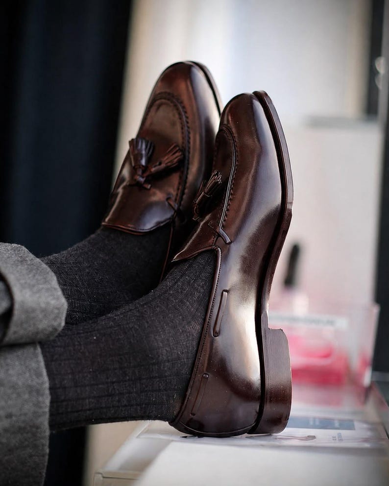 Puede incluir: Un par de mocasines de cuero marr&oacute;n oscuro con borlas y acabado pulido, con puntera cosida estilo mocas&iacute;n y tac&oacute;n bajo, usados con calcetines acanalados gris oscuro y pantalones grises, mostrando un dise&ntilde;o de zapato sin cordones cl&aacute;sico.
