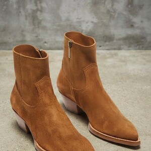 May include: A pair of tan suede ankle boots featuring a pointed toe, a stacked wooden heel, and a side zipper closure. The boots have a structured silhouette with visible stitching details along the vamp and a smooth, uniform texture throughout.