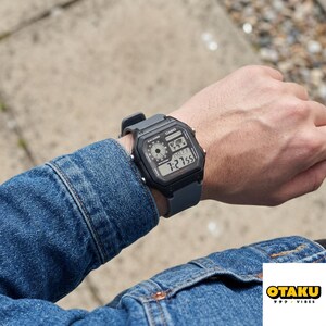 May include: A black digital watch with a gray band displays the time 7:27:55. The watch face includes a world map graphic and other digital displays. The watch is on a person's wrist, with a denim jacket in the background.