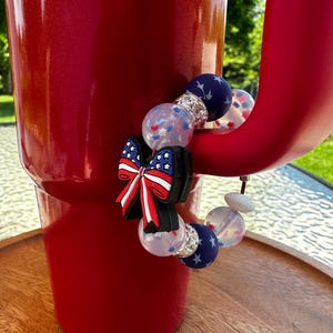 Puede incluir: Un vaso rojo con una pulsera decorativa. La pulsera presenta un lazo con un diseño de bandera estadounidense roja, blanca y azul, junto con cuentas transparentes y azul marino con acentos de estrellas. El vaso está sobre una superficie de madera.