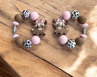 May include: Two brown and pink silicone bracelets with a cow charm. Each bracelet has brown, pink, and black and white patterned beads.