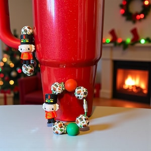May include: A large, red glittery tumbler with a handle, adorned with a charm bracelet featuring nutcracker and patterned ball charms. The bracelet is attached to the tumbler with magnets. Christmas-themed decor is visible in the background.