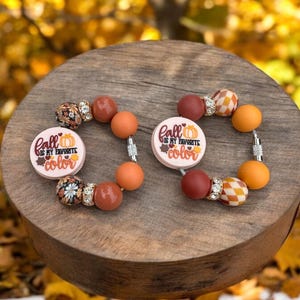 Puede incluir: Dos pulseras con temática otoñal con cuentas de color naranja, marrón y blanco. Una pulsera presenta un dije con el texto "el otoño es mi color favorito" y una calabaza. La otra pulsera presenta un dije con el texto "el otoño es mi color favorito" y una calabaza.