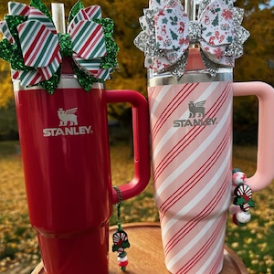 Peut inclure: Deux gobelets Stanley avec des nœuds sur le thème de Noël. L'un est rouge avec une poignée verte et un nœud à rayures vertes, rouges et blanches. L'autre est rose avec une poignée rose et un nœud à motifs blancs, rouges et verts. Les deux ont un charme.