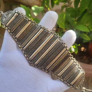 Può includere: Bracciale argentato dal design geometrico. Il bracciale presenta barre rettangolari con linee diagonali e motivi a puntini. I bordi sono collegati da una catena. Il bracciale è tenuto da una mano guantata su uno sfondo di fogliame verde.