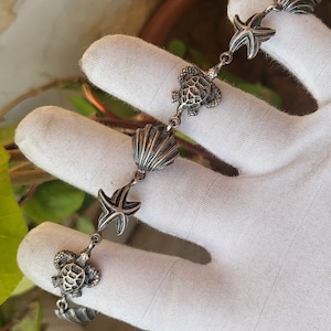 Può includere: Braccialetto d'argento con un design a tema marino. Il braccialetto presenta ciondoli a forma di conchiglia, stella marina e tartarughe marine.
