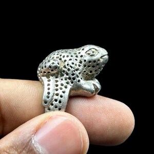 May include: A silver frog ring with a detailed, textured design. The frog is sitting on a finger.