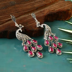 May include: A pair of silver peacock earrings with red gemstone accents. The earrings are detailed with intricate peacock designs and feature a dangling design.