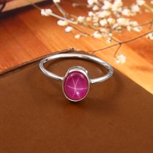 Może przedstawiać: Srebrny pierścionek z owalnym, gwiazdkowym kamieniem szlachetnym z rubinu. Rubin ma żywy różowy kolor, a pierścionek ma prosty, elegancki design. Pierścionek leży na brązowej powierzchni.