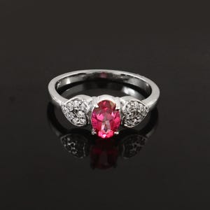 May include: A silver ring with a central oval-cut pink gemstone. The ring's band has small, clear accent stones on either side of the main stone. The ring is displayed against a dark background, highlighting its reflective qualities.