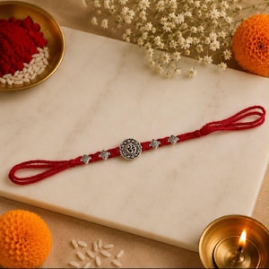 May include: A red thread bracelet featuring a central circular silver-toned charm embossed with the Om symbol, flanked by four smaller silver-toned decorative beads. The bracelet has looped ends and is constructed from braided red cord material.