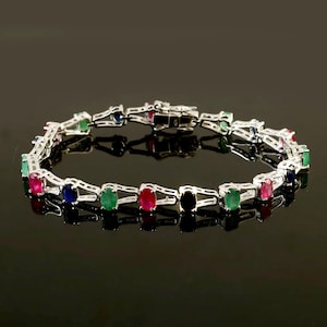 May include: A silver bracelet with a row of oval-shaped gemstones in red, green, blue, and black. The bracelet has a clasp closure.