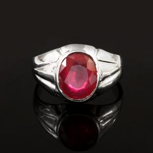 Può includere: Un anello d'argento con una grande gemma di rubino taglio ovale. Il rubino è di colore rosso intenso ed è incastonato in una fascia d'argento lucida. L'anello è esposto su una superficie nera riflettente.