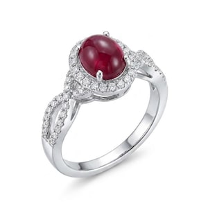 May include: A silver ring featuring a large, oval-cut ruby gemstone surrounded by a halo of smaller, clear gemstones. The ring's band has a twisted design, also adorned with small, clear gemstones, creating a sparkling and elegant appearance.