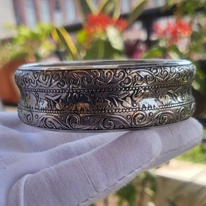 May include: A silver bangle bracelet with an intricate design of swirling floral patterns and small elephant figures. The bracelet is made of metal and has a polished finish.
