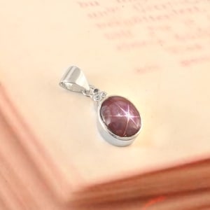 Op de afbeelding: Een zilveren hanger met een ovale, stervormige robijn edelsteen. De robijn heeft een diepe rood-paarse kleur en de hanger heeft een eenvoudig, elegant ontwerp. De hanger is waarschijnlijk gemaakt van sterling zilver.