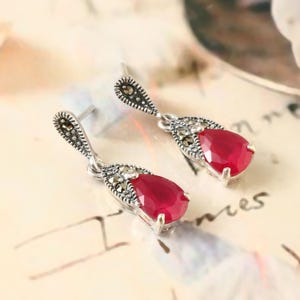 May include: A pair of silver dangle earrings with red pear-shaped gemstones and marcasite accents.