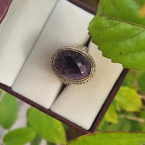 Könnte beinhalten: Ein silberner Ring mit einem großen, ovalen Amethyst-Edelstein. Der Edelstein ist facettiert und hat eine tiefviolette Farbe. Der Ring ist in einer silbernen Band mit einem dekorativen, gedrehten Design gefasst.