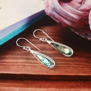 May include: Silver teardrop earrings featuring iridescent abalone shell inlays. The earrings have a hook closure and a polished finish. The abalone shell displays shades of blue, green, and purple. The earrings are displayed on a wooden surface.
