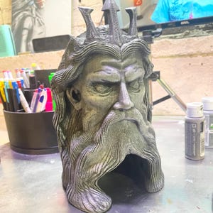 May include: A grey sculpture of a bearded man wearing a crown and holding a trident. The sculpture has long hair and a detailed beard. The sculpture is made of a stone-like material and has a weathered appearance. The sculpture is a bust of a mythological figure.