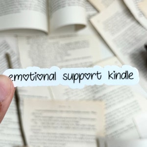 May include: White sticker with the text "emotional support kindle" with a heart symbol in place of the "o" in emotional and support.