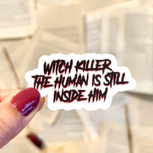 May include: A white sticker with the text "WITCH KILLER THE HUMAN IS STILL INSIDE HIM" in black and red. The sticker is held by a hand with a red painted nail, with a background of open book pages.