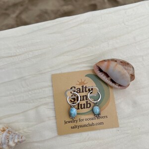 May include: Silver hoop earrings with oval, light blue gemstones. The earrings are displayed on a card with the text "Salty Sun Club" and "Jewelry for ocean lovers". A seashell is next to the earrings.