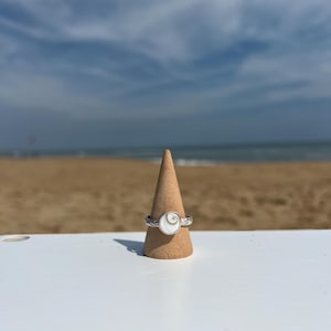 May include: A silver ring with a white, circular centerpiece and a twisted band is displayed on a wooden cone. The ring is set against a backdrop of a beach, ocean, and sky.