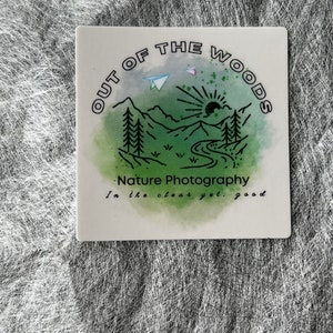 May include: Square white tile featuring a nature-themed design. The design includes the text "OUT OF THE WOODS" and "Nature Photography" with a watercolour illustration of mountains, trees, a river, and a sun. The phrase "In the clear yet, good" is also included.