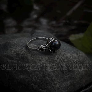 May include: A silver ring with a black pearl set in an intricate design. The ring is resting on a dark grey stone.