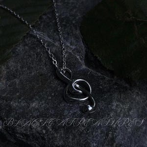 Puede incluir: Un collar plateado con un colgante de clave de sol. El colgante cuelga de una cadena delicada. El collar se muestra sobre una superficie oscura y texturizada. El texto "BLACK TREASURES" está en la parte inferior de la imagen.