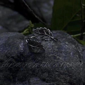 Pode incluir: Um anel prateado com um design de folha. O anel apresenta duas formas de folhas detalhadas que envolvem o dedo. O anel está sobre um fundo escuro e texturizado. O texto "BLACKTREASURES" está na parte inferior da imagem.