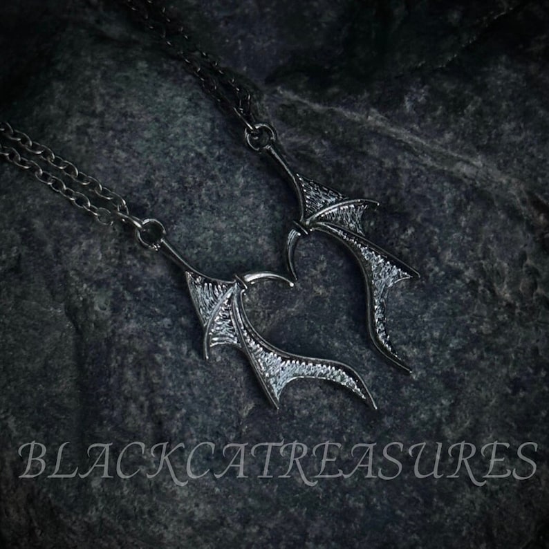 May include: Two silver necklaces with detailed, intricate, wing-shaped pendants. The wings are facing each other, creating a heart shape. The necklaces are on a dark, textured background.