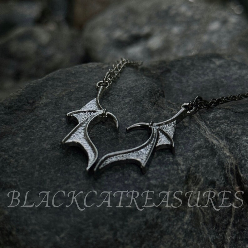 May include: A silver necklace with a pendant shaped like two intertwined wings. The wings are detailed with a textured pattern and have a slightly gothic aesthetic.