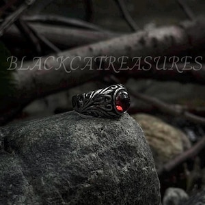 May include: A silver ring with an ornate design and a red gemstone. The ring is set on a gray rock.