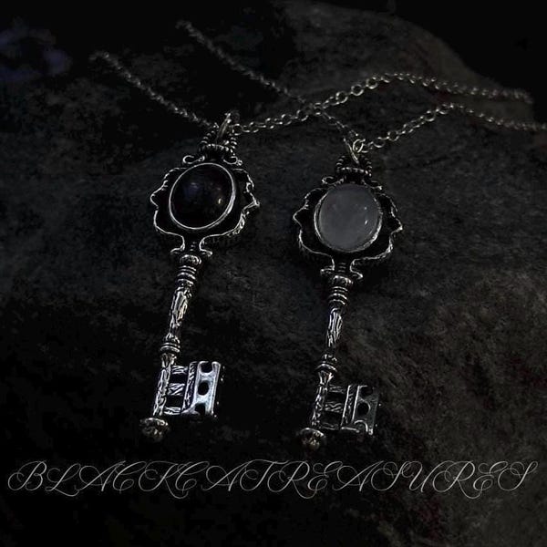 May include: Two ornate key pendant necklaces. One key features a dark stone, the other a light stone, both set in decorative silver-toned frames. The keys hang from delicate chains, with the words "BLACK TREASURES" at the bottom.