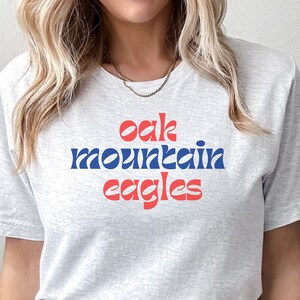 May include: A light gray t-shirt with the words "Oak Mountain Eagles" in red and blue lettering. The shirt is made of a soft material and has a crew neck.