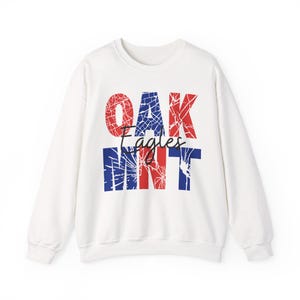 May include: White crew neck sweatshirt featuring the text "OAK Eagles MINT" in red and blue. The text has a distressed, cracked design. The sweatshirt has long sleeves.